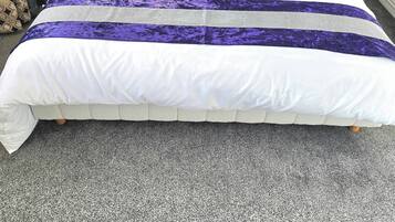 Un lit bien fait, recouvert d’une couvre-lit blanche et violette, avec une tête de lit ornée d’un motif floral et un tapis gris.