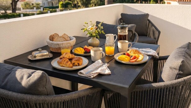 Une table dressée avec des aliments pour le petit-déjeuner, une cafetière et un pichet de jus d’orange, sur une terrasse extérieure agrémentée de chaises en osier et d’un palmier en arrière-plan.