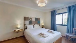 Une chambre d’hôtel avec un grand lit, deux lampes de chevet, une tête de lit ornée d’un motif en patchwork, une chaise et une fenêtre avec des rideaux bleus.