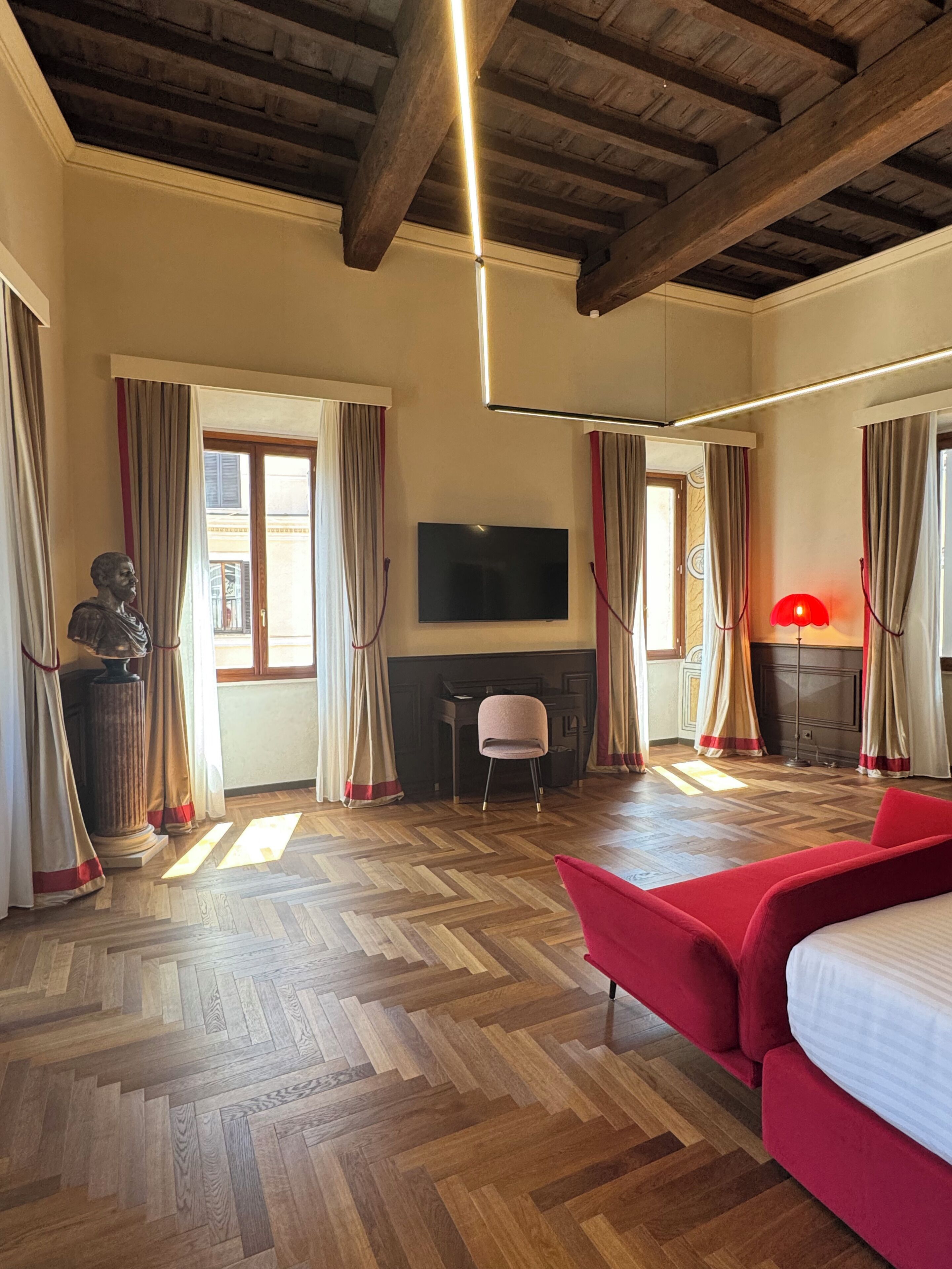 A hotel room with a wooden floor, a red sofa, a television, a fireplace, and a large window with curtains.
