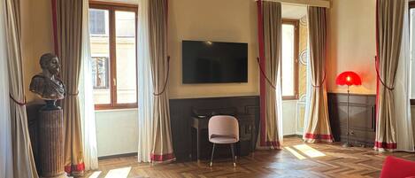 A hotel room with a wooden floor, a red sofa, a television, a fireplace, and a large window with curtains.
