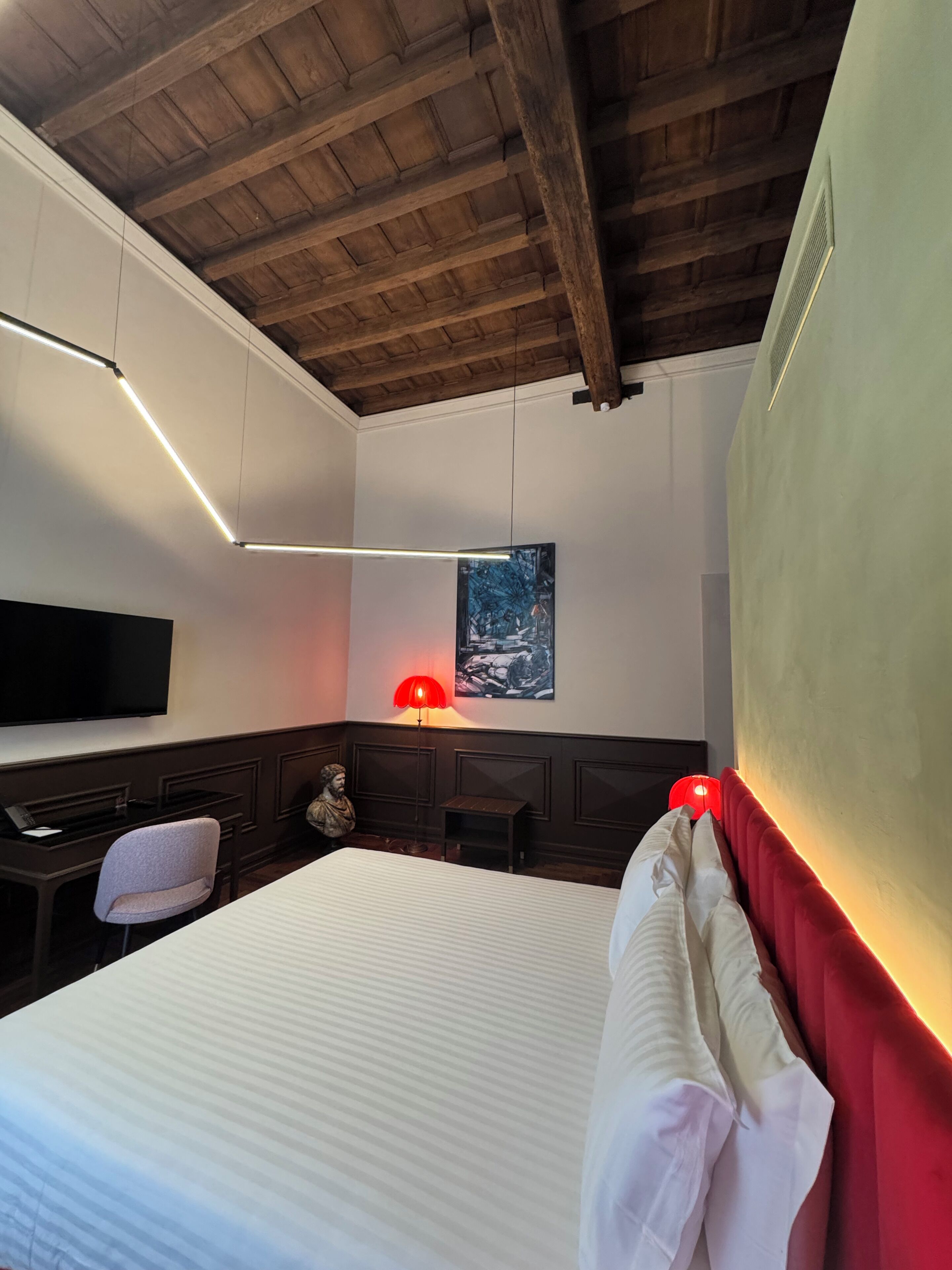 A hotel room with a wooden ceiling, a bed with white bedding, a desk with a chair, a television, and a red lamp.