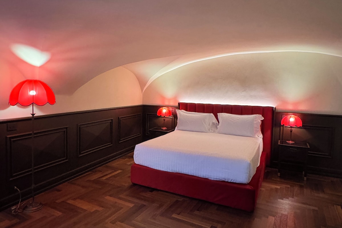 A bedroom with a red bed, two bedside lamps, a wooden herringbone floor, and a curved ceiling.