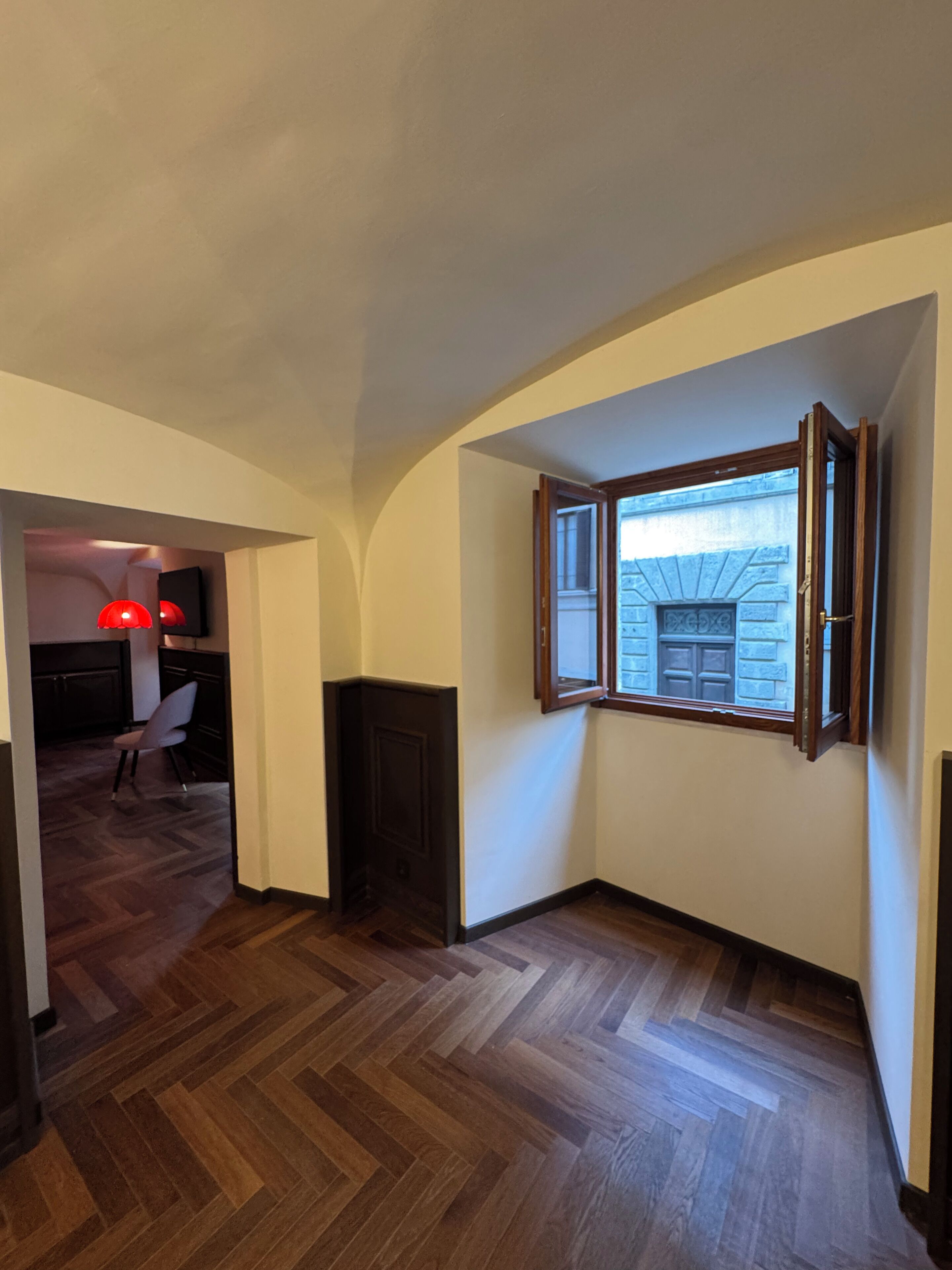 Un pasillo con piso de madera, una puerta y un ventanal con contraventanas abiertas.