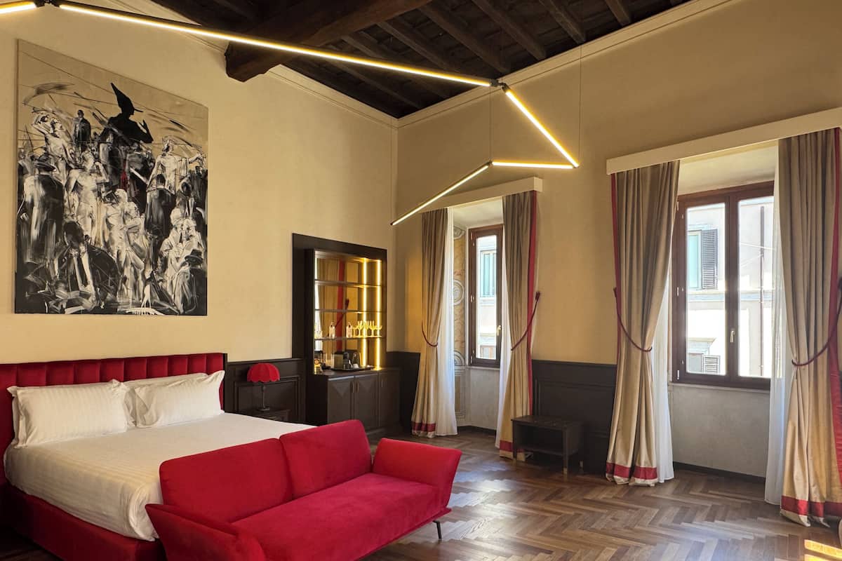 A hotel room with a bed, red sofa, wooden parquet floor, a painting on the wall, a mirror, and a window with curtains.