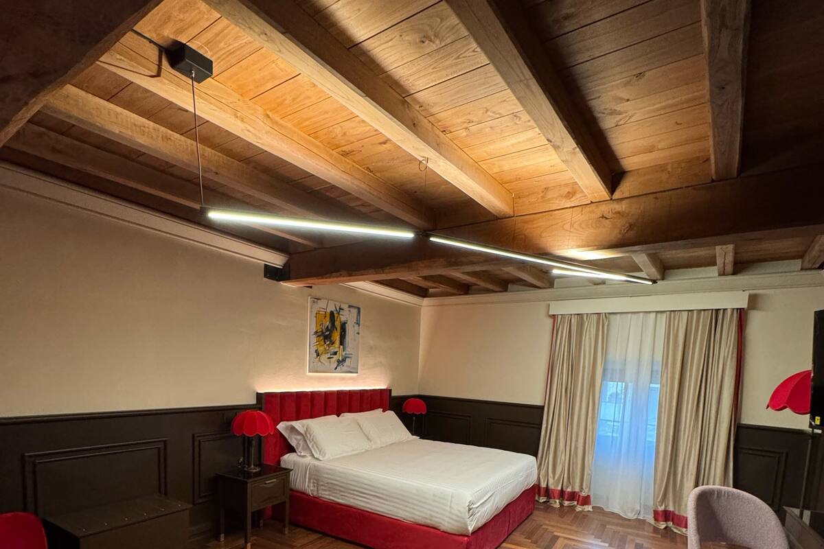 A bedroom with a wooden ceiling, a bed, a red chair, a bedside table, and a window with curtains.