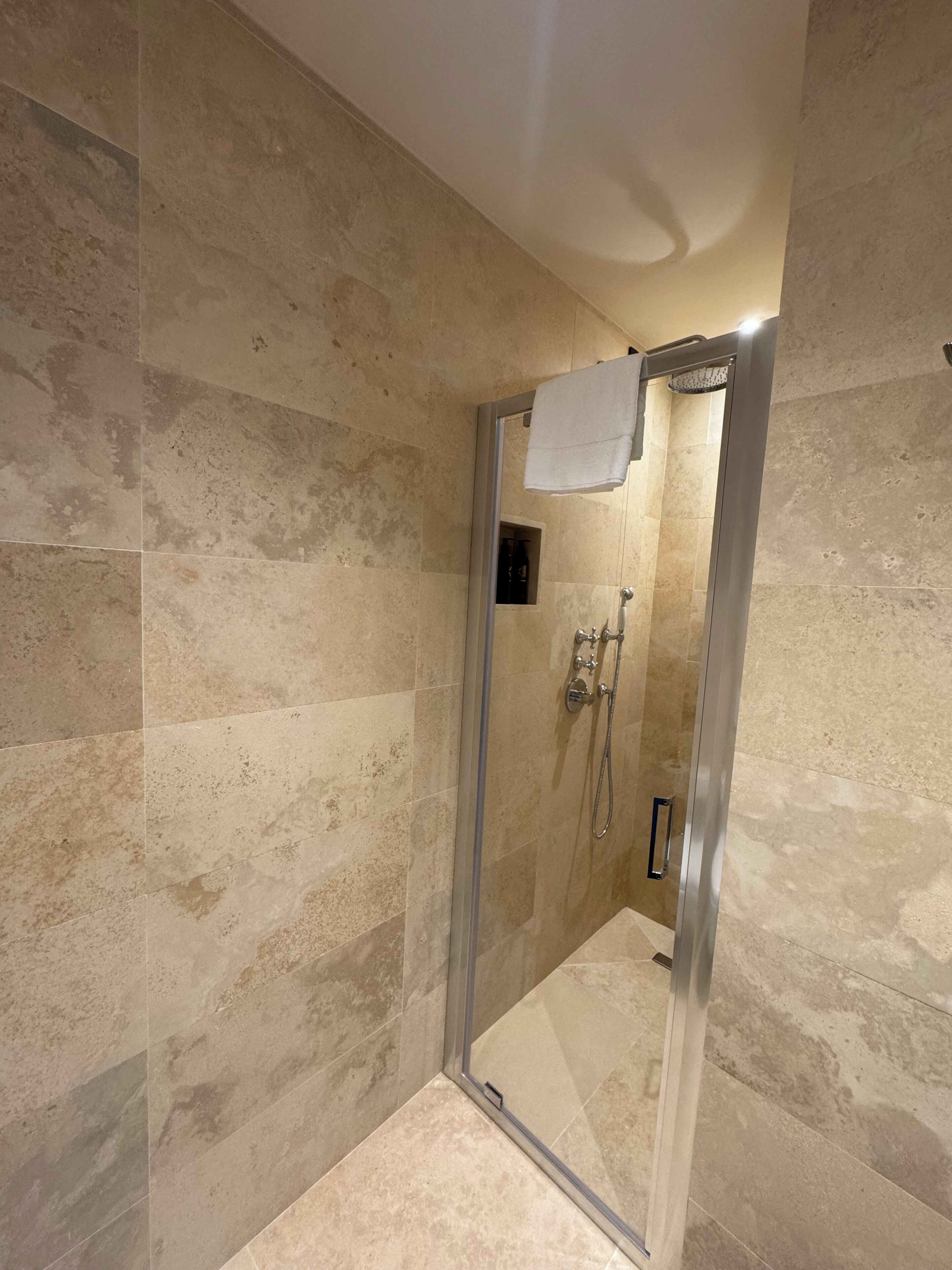 A glass-enclosed shower with beige tiles, a towel hanging on the door, and a showerhead.