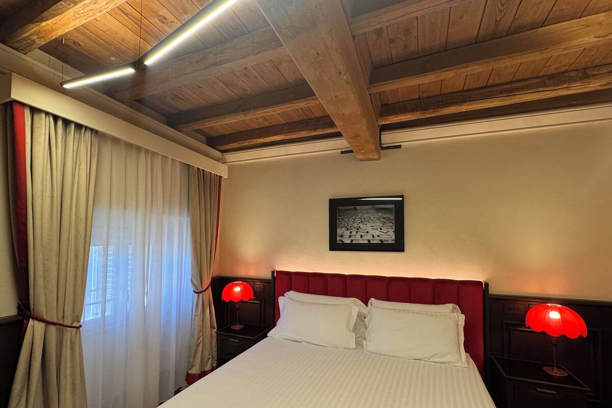 A hotel room with a wooden ceiling, a large bed with white bedding, two bedside tables with red lamps, and a framed picture on the wall.