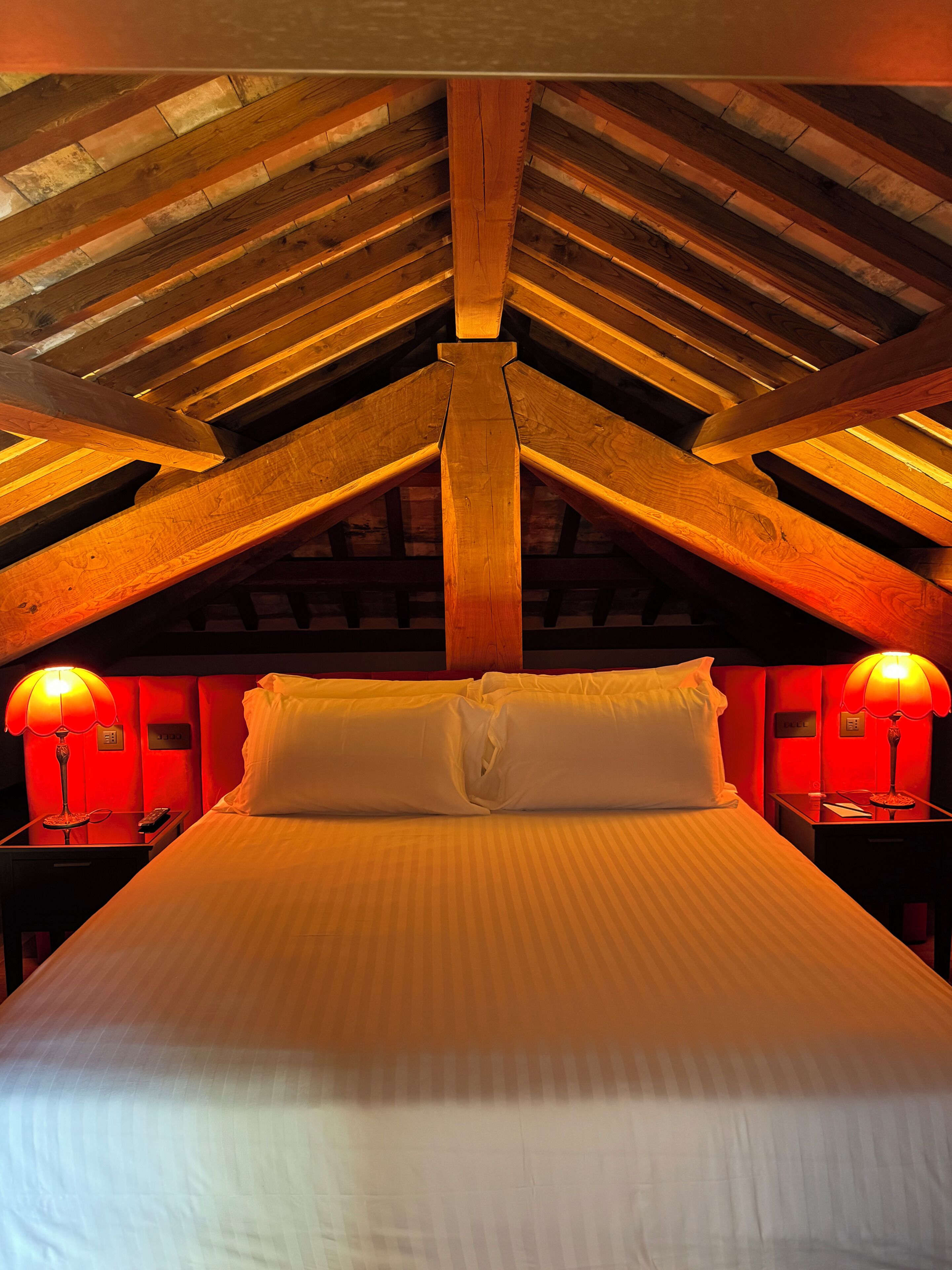 Een hotelkamer met een hoge plafond, houten balken, een bed met witte lakens, twee nachtlampjes en een rode accentmuur.