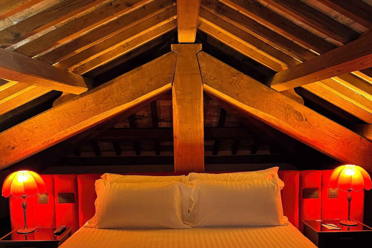 A hotel room with a high ceiling, wooden beams, a bed with white linens, two bedside lamps, and a red accent wall.