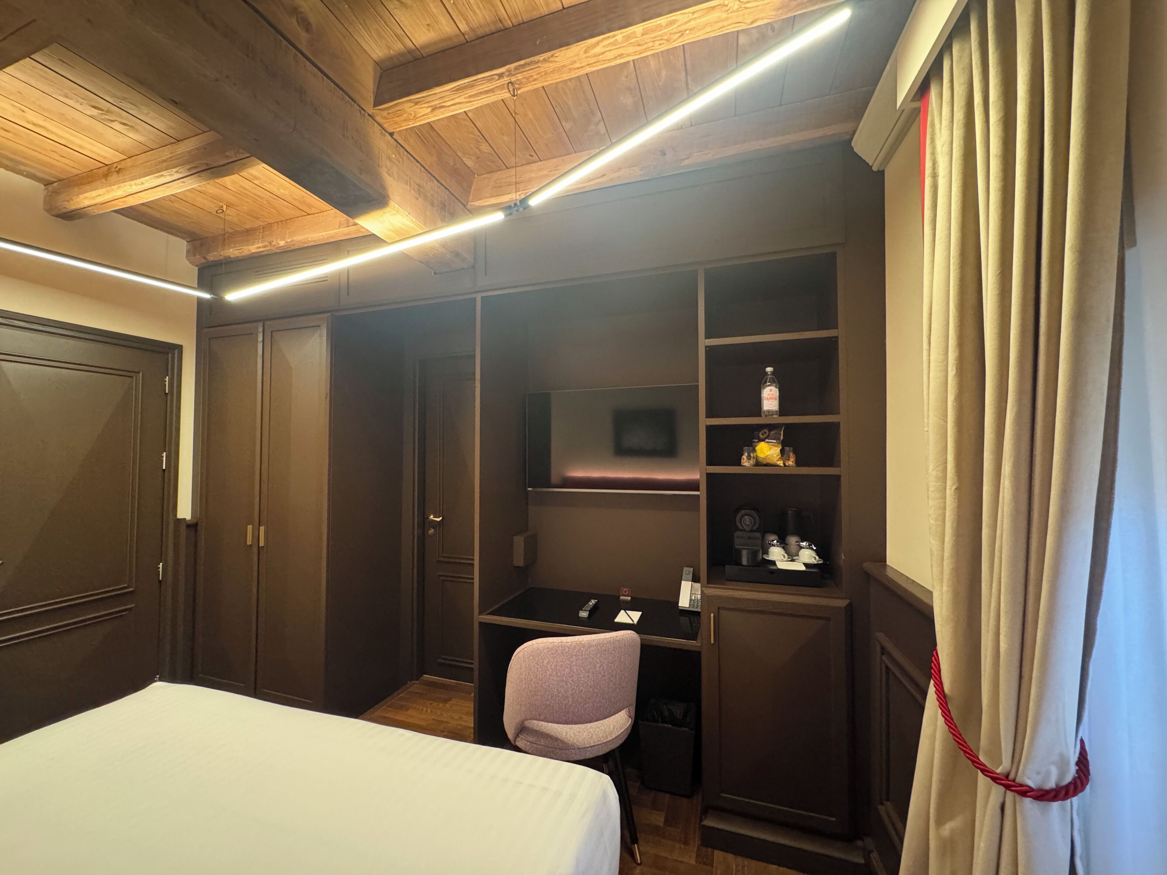A hotel room with a bed, a desk, a chair, a wardrobe, and a shelf with toiletries.
