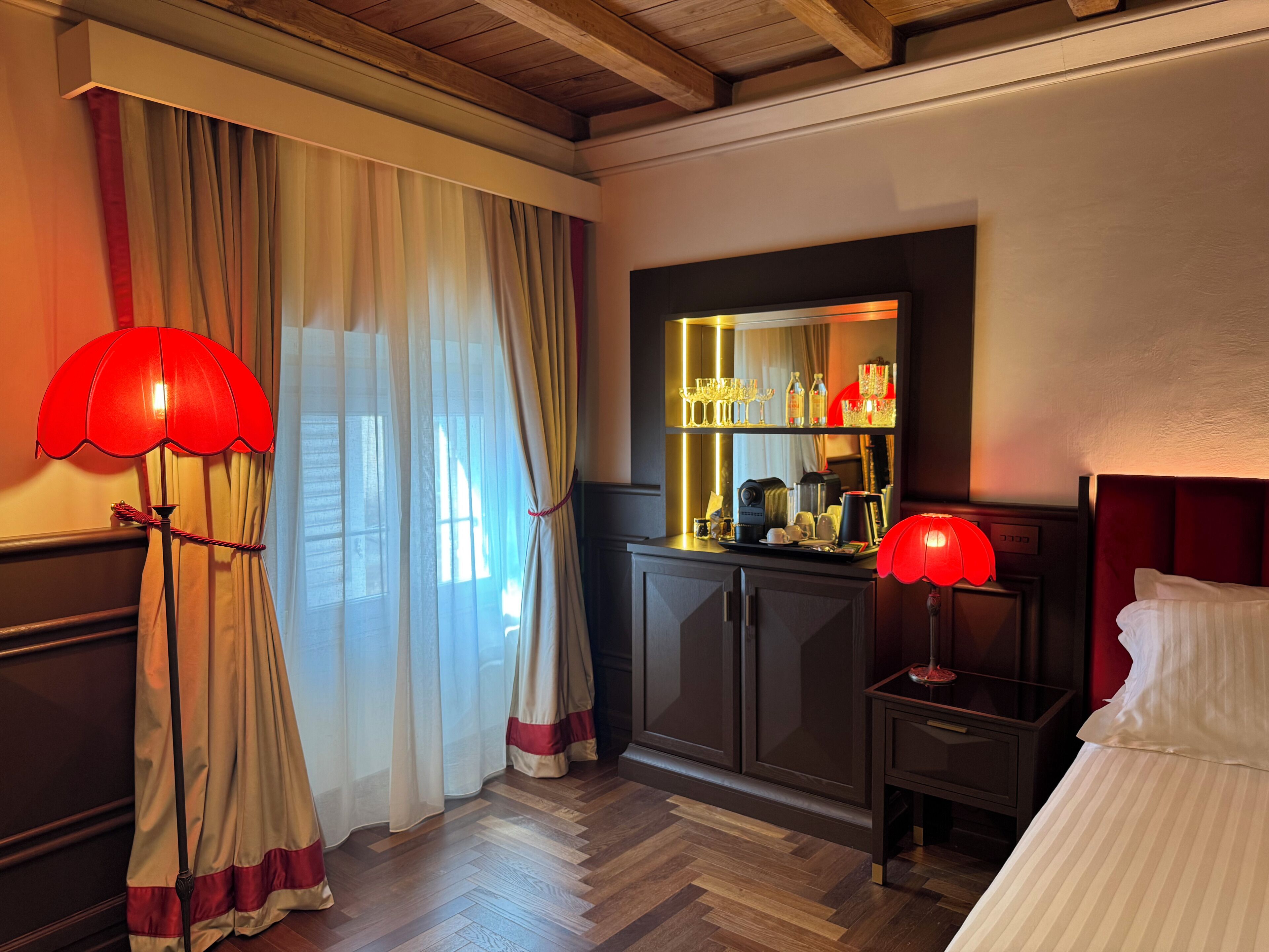 Habitación de hotel con piso de madera, una cama, una mesita de noche, una lámpara roja, un espejo y una ventana con cortinas.