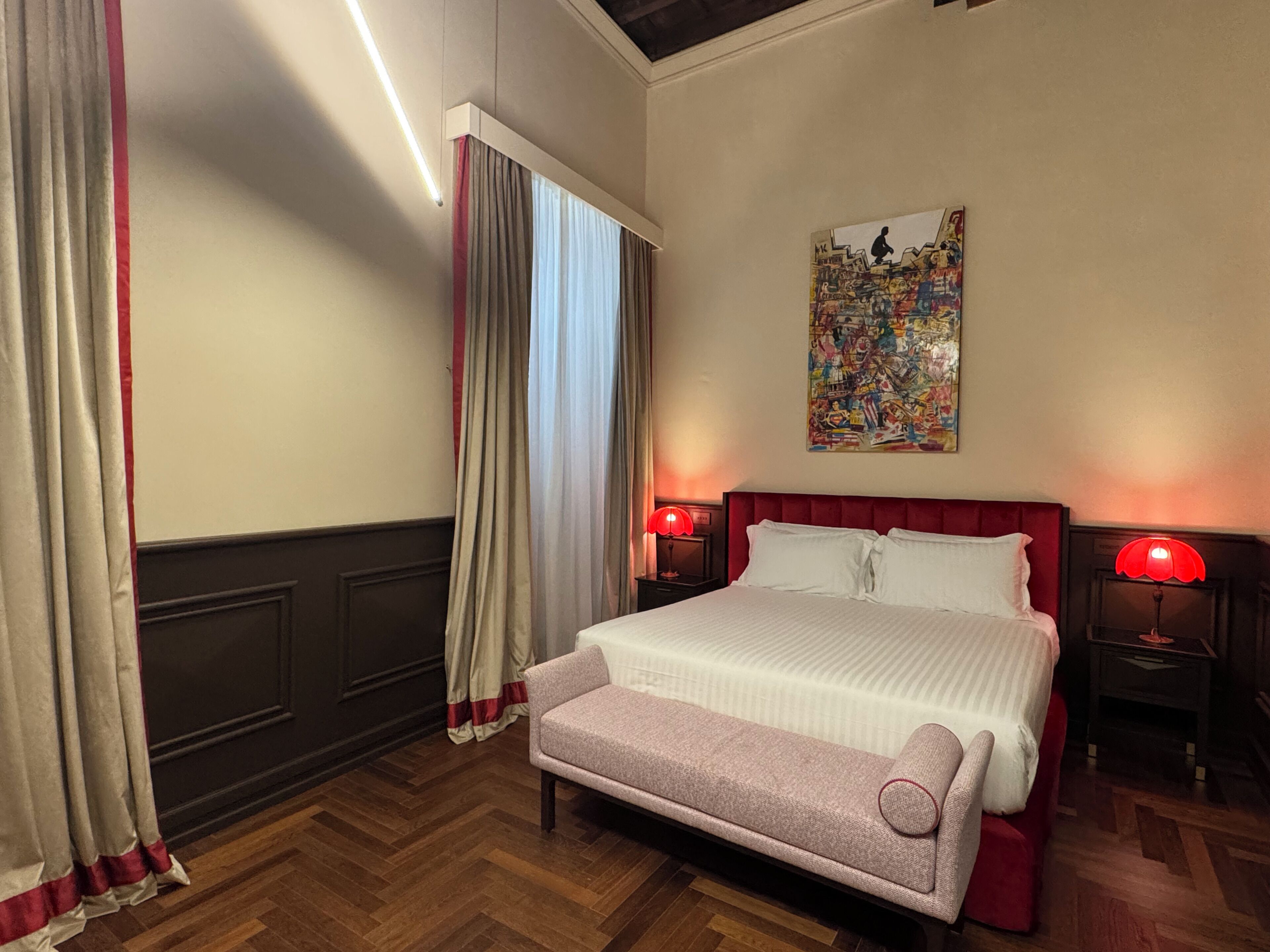 A hotel room with a large bed, two bedside tables with lamps, a bench, a window with curtains, and a wall with a colorful painting.