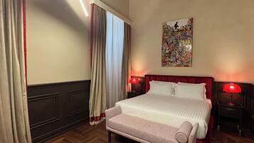 A hotel room with a large bed, two bedside tables with lamps, a bench, a window with curtains, and a wall with a colorful painting.