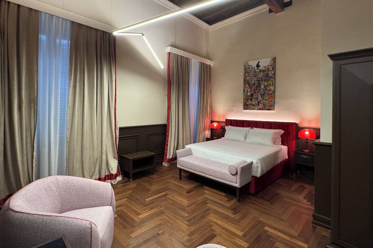 A bedroom with a bed, two bedside tables with lamps, a chair, a wooden floor, and a painting on the wall.