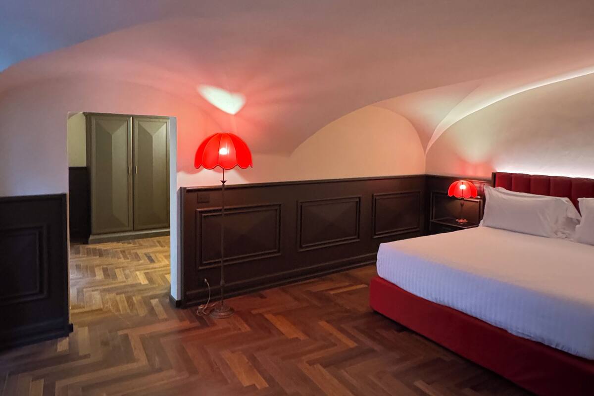 A bedroom with a wooden herringbone floor, a bed with white bedding, a red lamp, and a dark headboard.