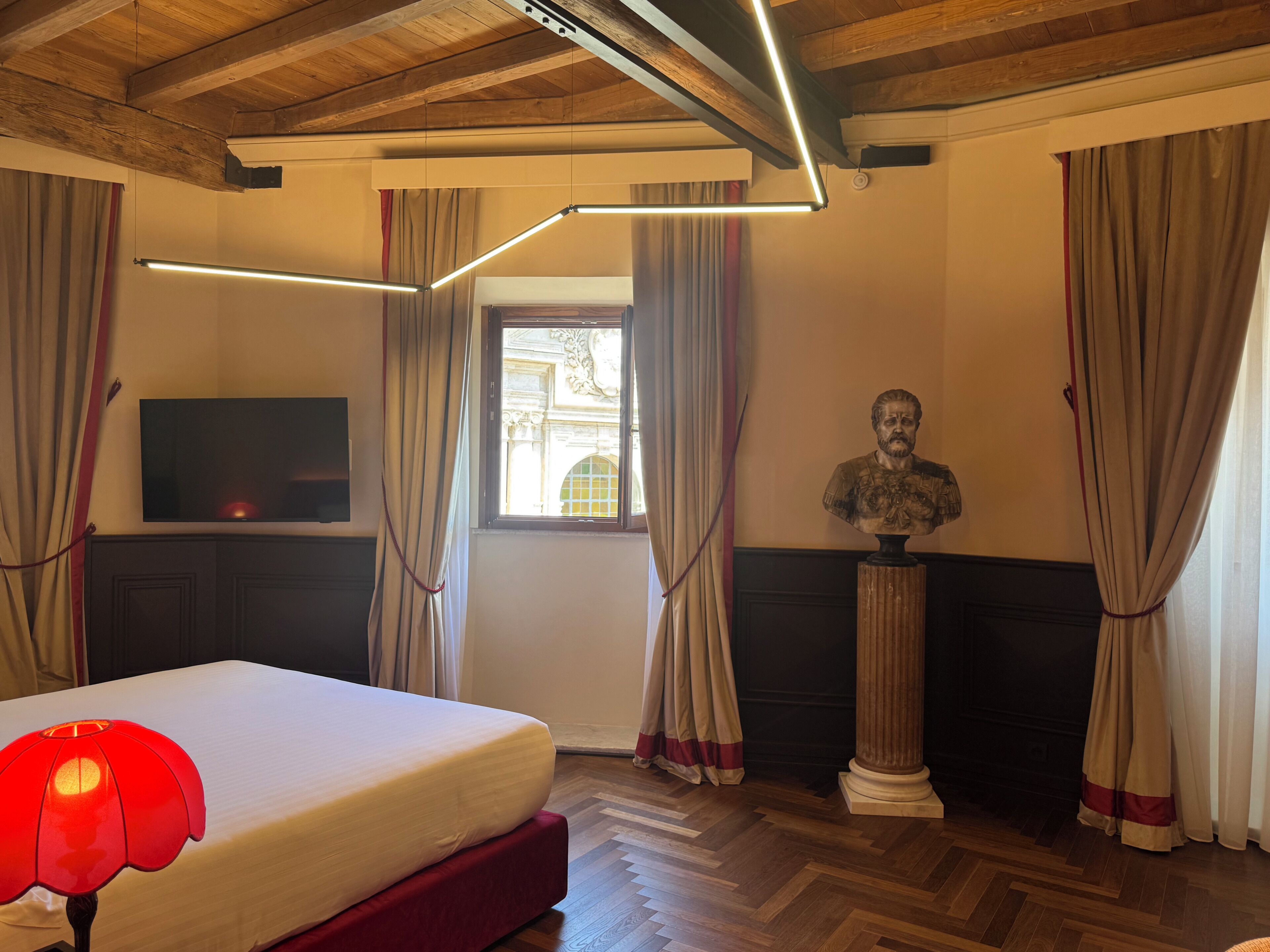A room with a bed, a flat-screen TV, a bust sculpture, and a window with curtains.