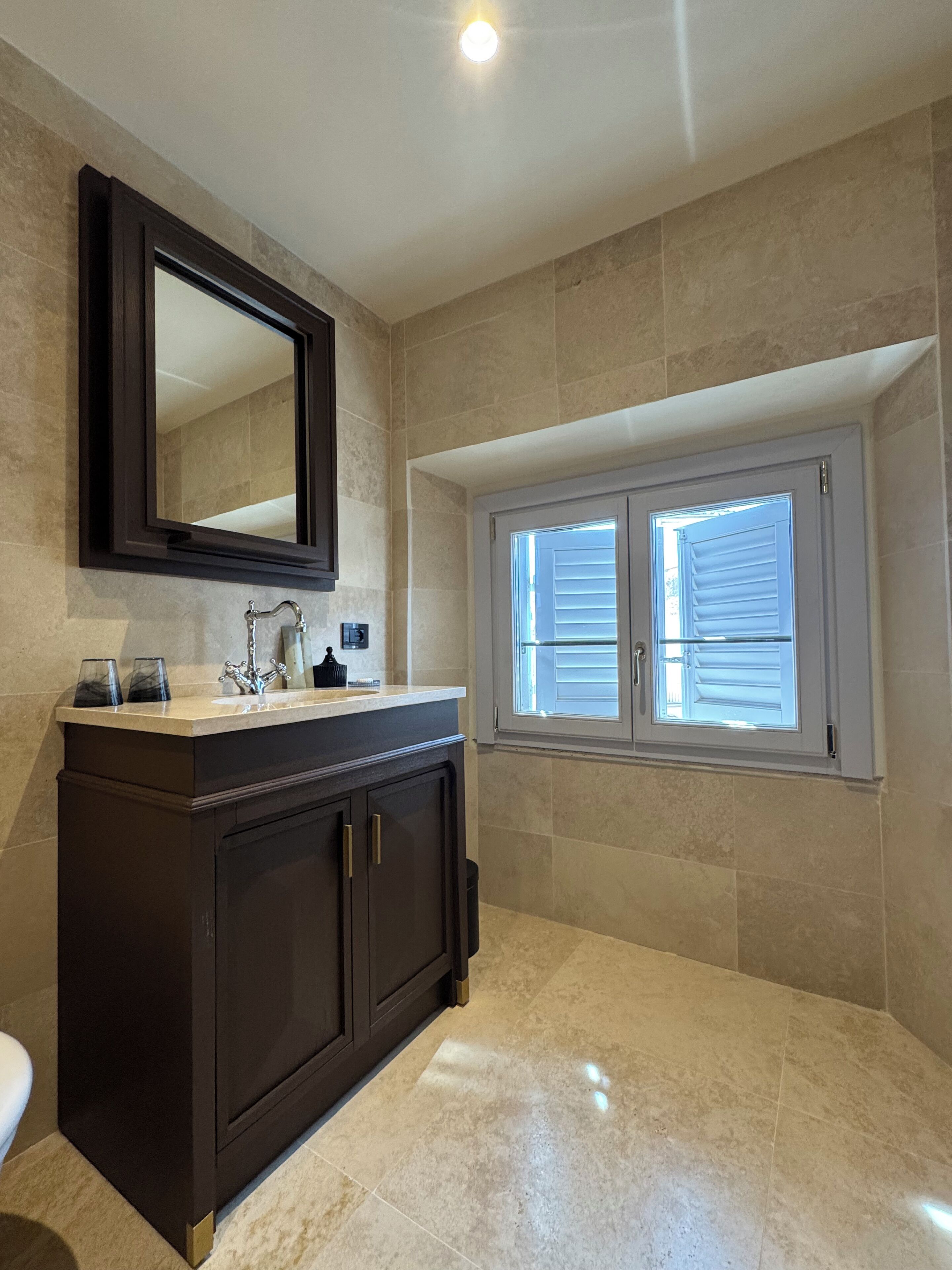 A bathroom with a dark vanity, a large mirror, a window with blinds, and a light fixture on the ceiling.