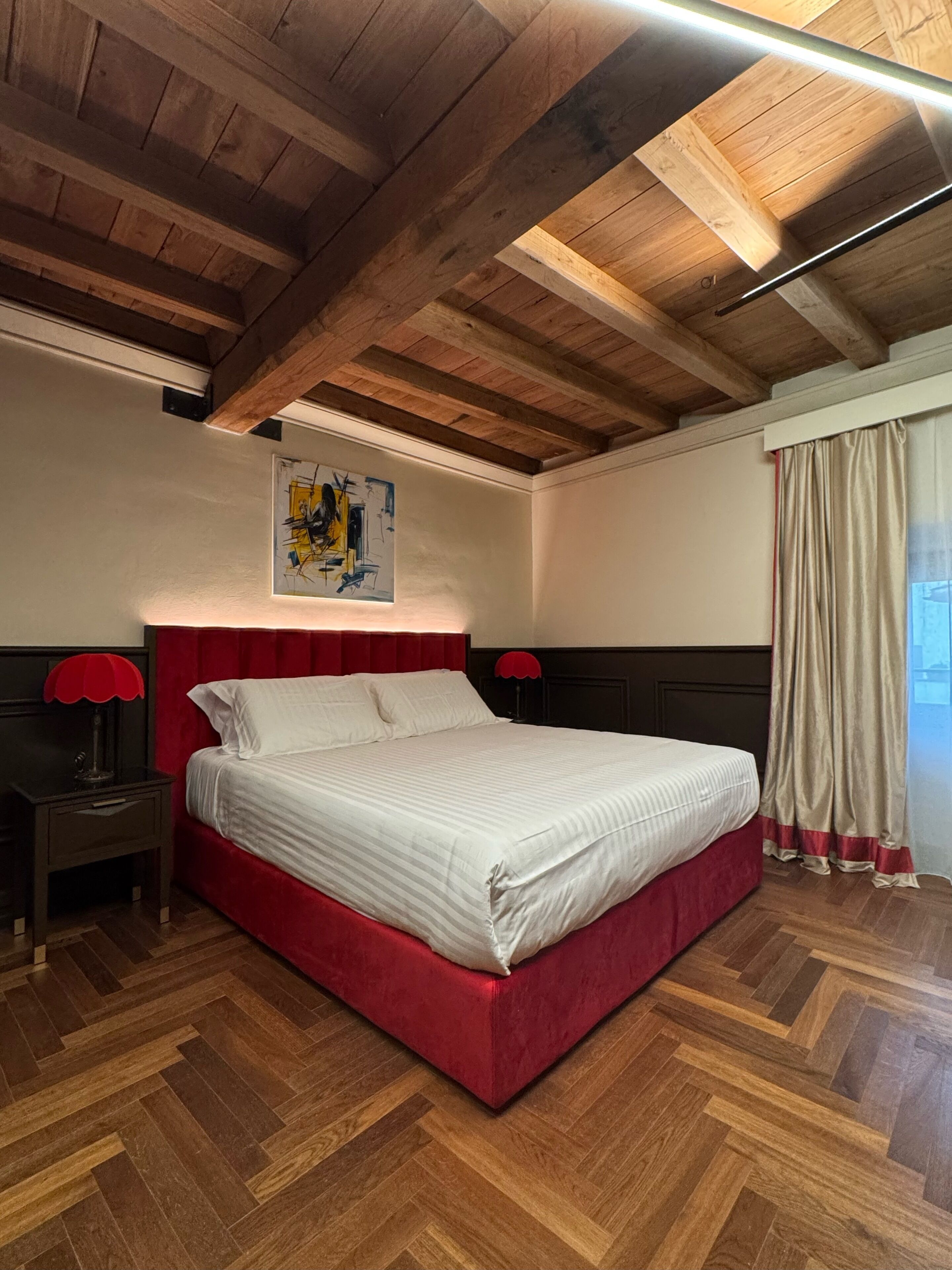 Un dormitori amb sostre de fusta, un llit amb capçalera tapissada de color vermell, una tauleta de nit amb una làmpada i un quadre a la paret.