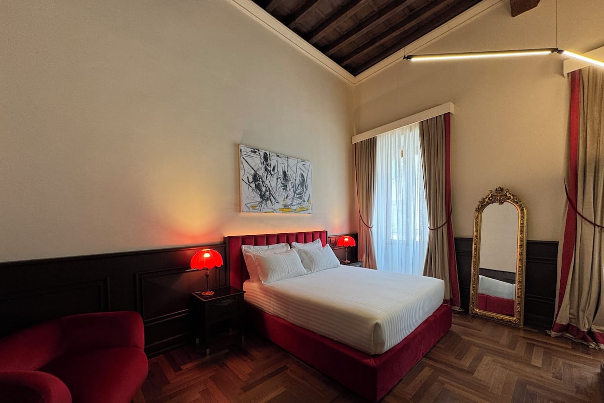 A hotel room with a bed, two bedside tables with lamps, a red chair, a large mirror, and a painting on the wall.