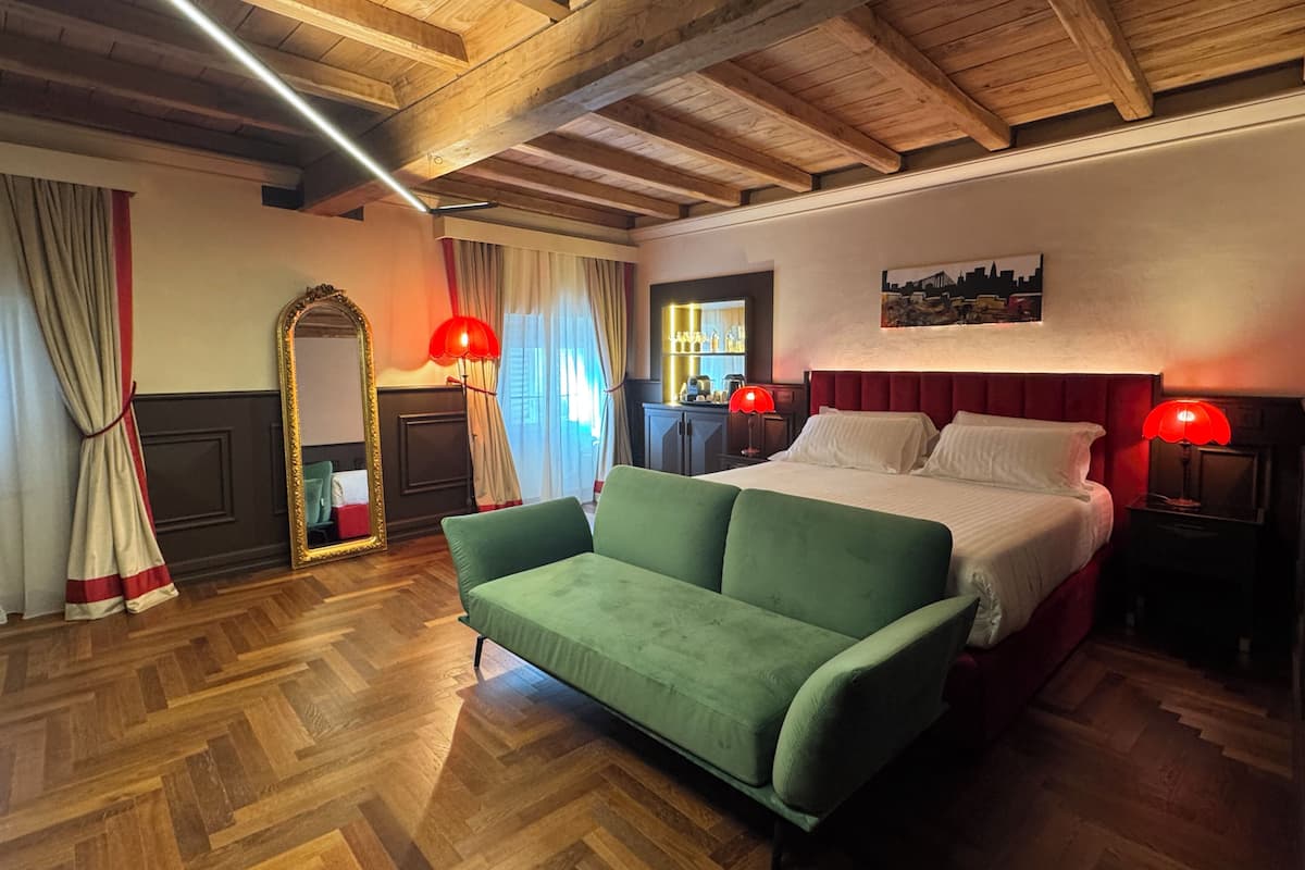 A hotel room with a wooden ceiling, a green sofa, a bed with white linens, red lamps, and a cityscape painting.