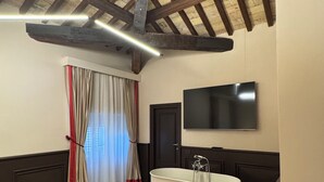Un baño con bañera, vigas de madera en el techo, un televisor de pantalla plana y una ventana con cortinas.