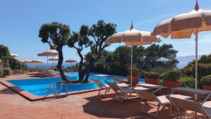 Ein Poolbereich mit Liegestühlen und Sonnenschirmen, umgeben von Bäumen und Blick aufs Meer.