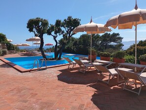 Ein Poolbereich mit Liegestühlen und Sonnenschirmen, umgeben von Bäumen und Blick aufs Meer.