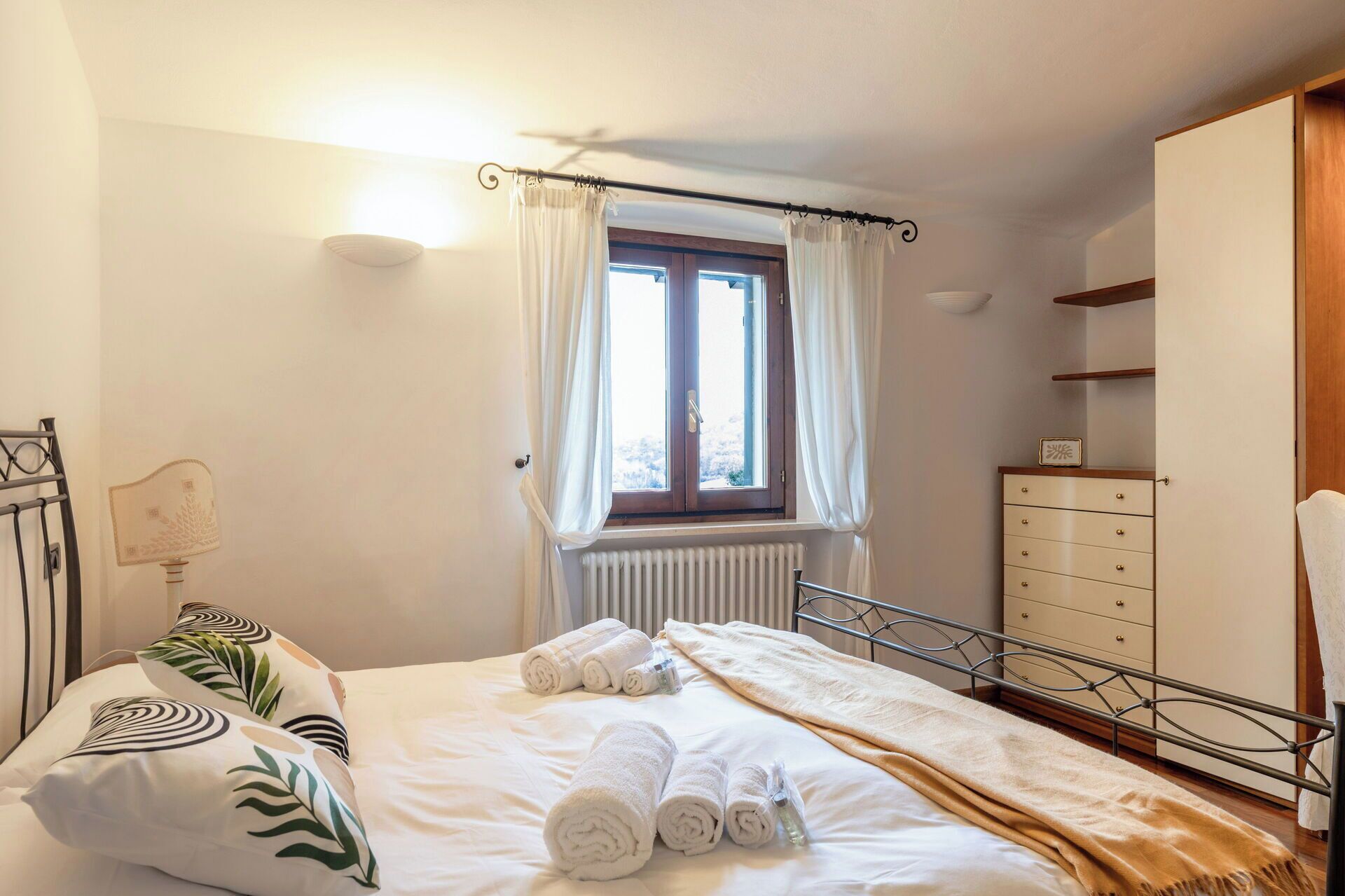 A bedroom with a bed, a radiator, a window with curtains, a bedside table, and a wardrobe.