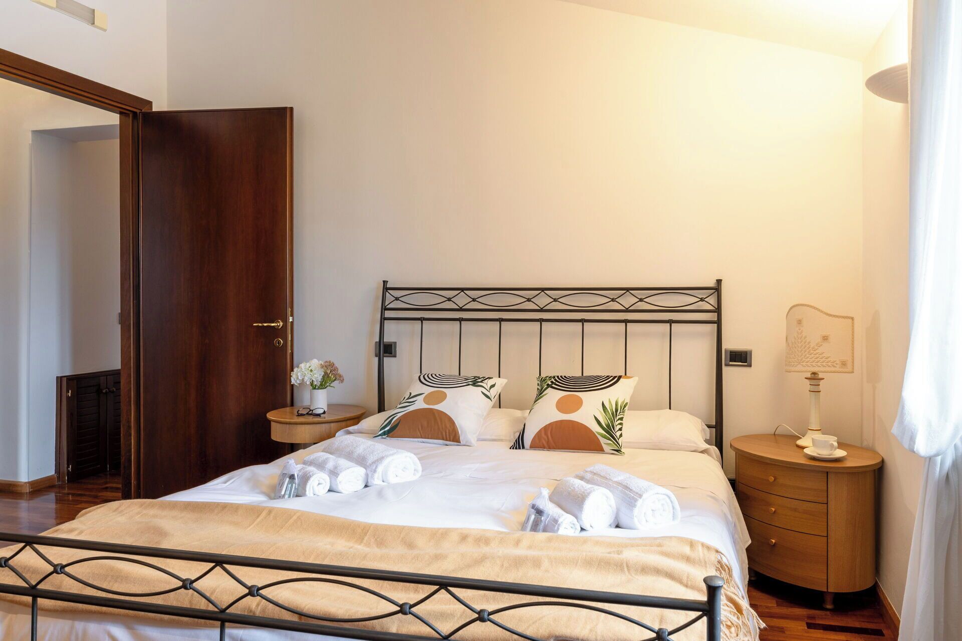 Ein ordentlich bezogenes Bett mit weißen Leinen und aufgerollten Handtüchern, ein Nachttisch aus Holz mit einer Lampe sowie ein an der Wand befestigter Bilderrahmen.