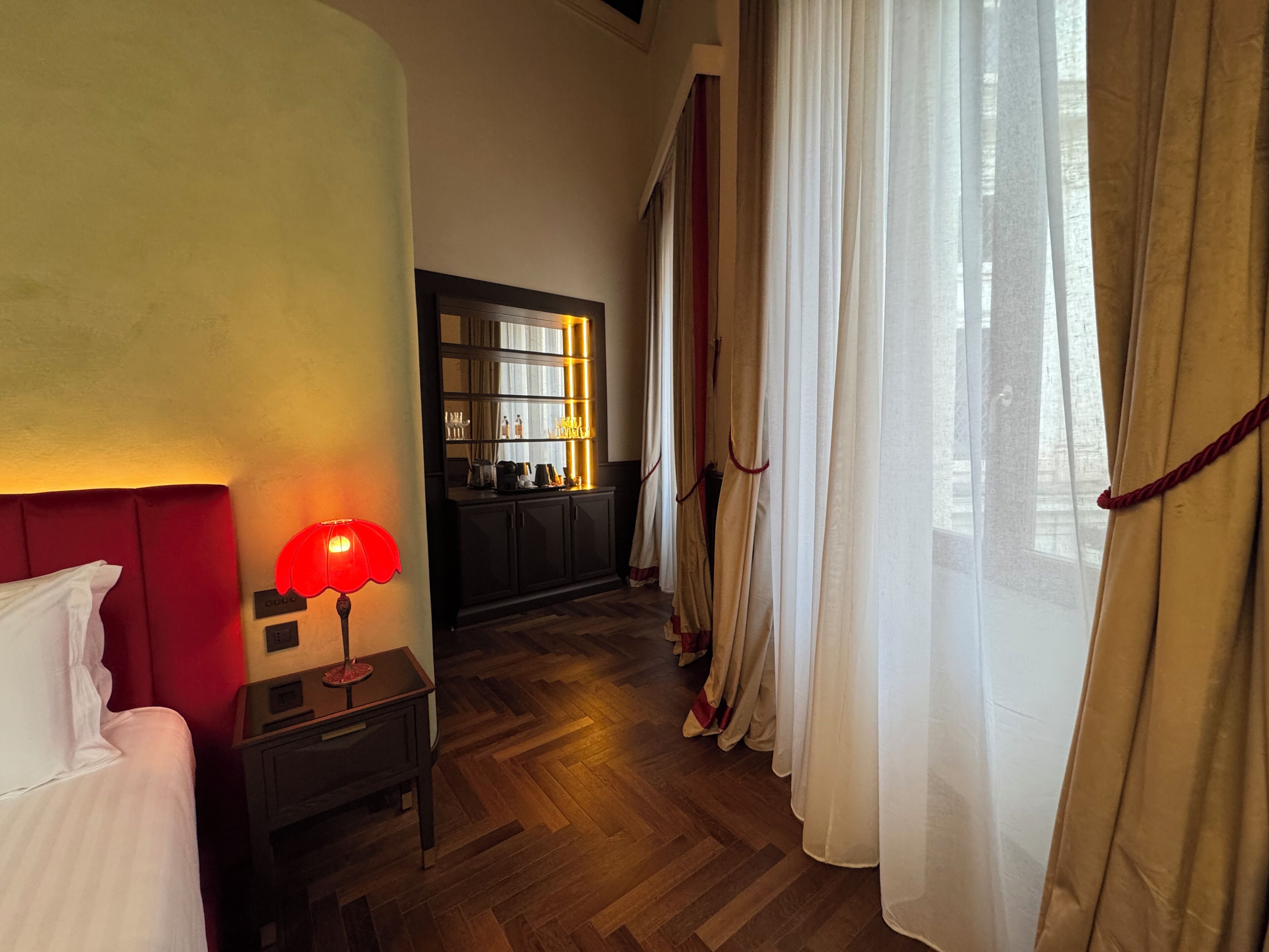 A hotel room with a wooden floor, a bed with white bedding, a nightstand with a red lamp, a mirror, a cabinet with items, and sheer curtains.