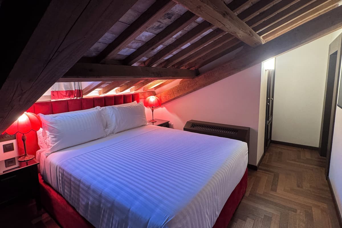 Ein Schlafzimmer im Dachgeschoss mit schräg angesetzten Decken, einem Bett mit weißen Leinen, roten Kissen und zwei roten Lampen.