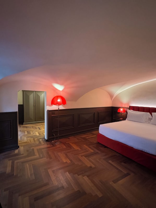 A bedroom with a wooden herringbone floor, a bed with white bedding, a red lamp, and a dark headboard. - Palazzo Gambirasi Aparthotel Roma (Rome)