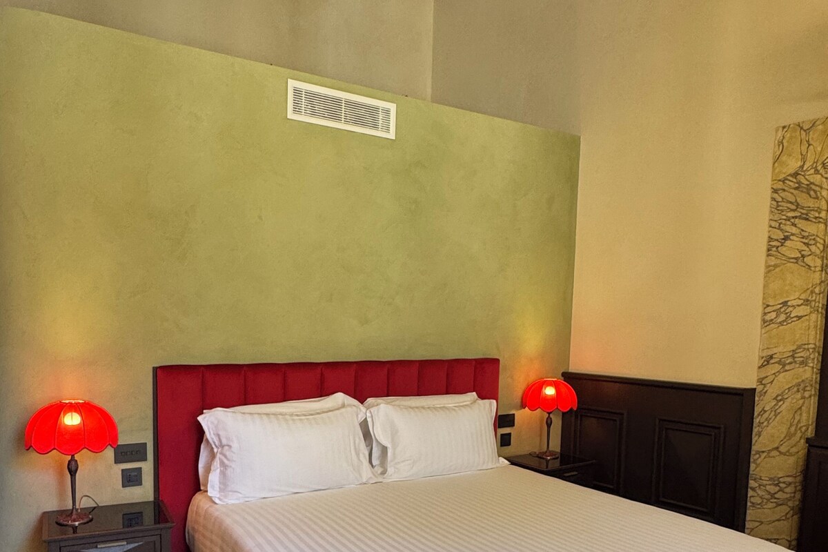 A bed with a red headboard, two bedside tables with lamps, a wooden floor, and a wall with a vent.