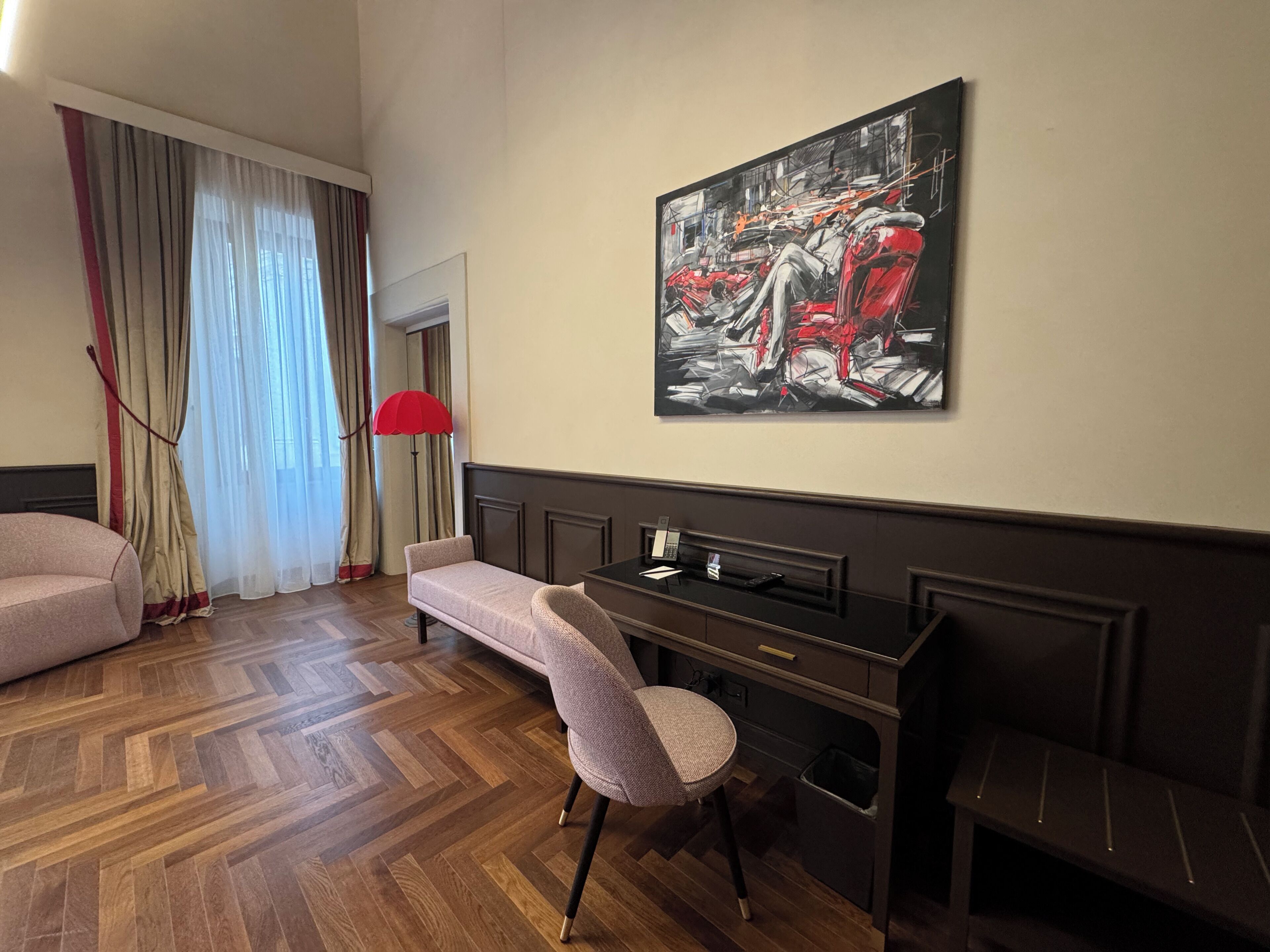 Ein Hotelzimmer mit einer Couch, einem Schreibtisch, einem Stuhl, einer roten Stehlampe und einem großen abstrakten Gemälde an der Wand.