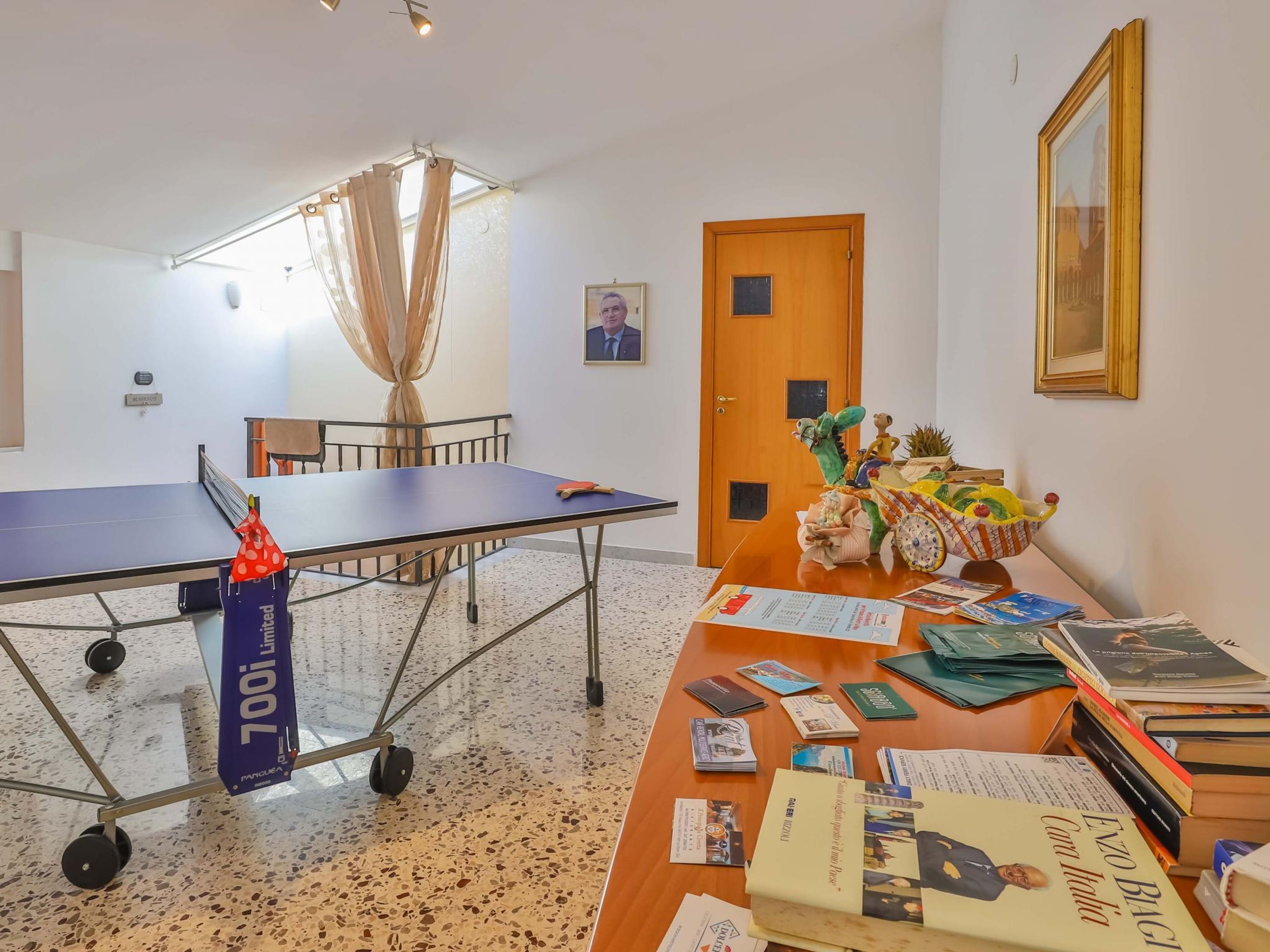 Une pièce avec une table de ping-pong, un bureau avec des livres et des magazines, une porte et un tableau encadré au mur.