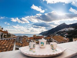 A terrace with a ceramic tray holding three cups and a teapot, overlooking a coastal town with mountains in the background.