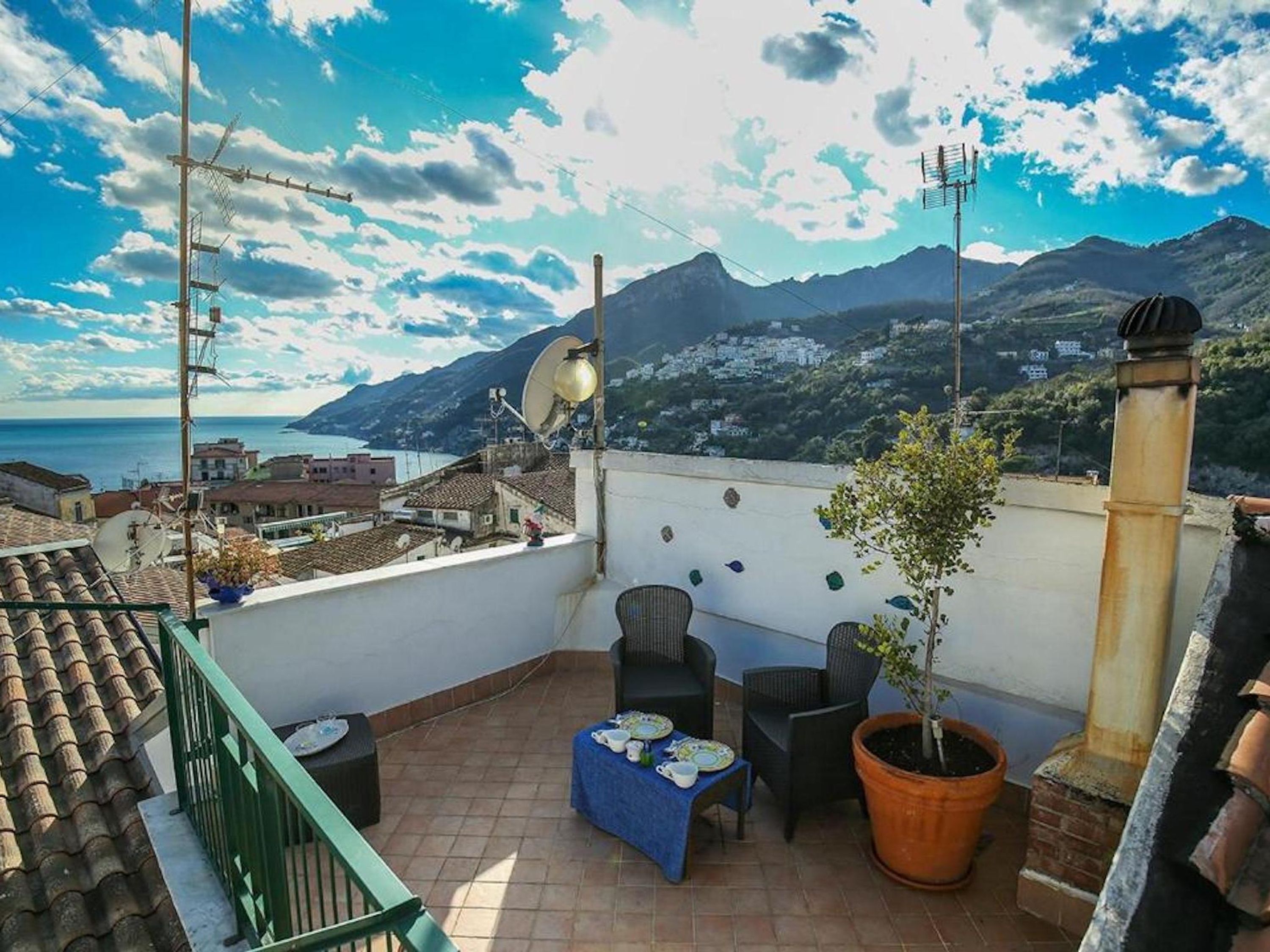 A rooftop terrace with a view of the sea and mountains, featuring outdoor furniture and a potted plant.