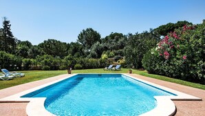 Ein rechteckiger Pool mit weißem Rand, umgeben von einer Pflasterterrasse. Im Hintergrund sind Liegestühle und Kübelpflanzen zu sehen.