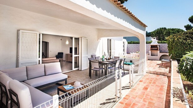 Eine überdachte Terrasse im Freien mit einer Couch, einem Esstisch, Stühlen, einem Grill und Blick auf die umliegende Landschaft.
