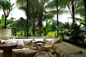 A room with a large window overlooking a lush garden, a bed with a view, and a small table with glasses. - Dzen Greenfield Villa 4 (Ubud)