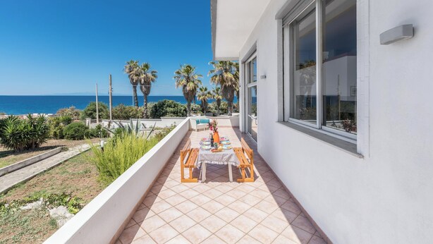 A balcony with a table and chairs, overlooking a pool and palm trees, with a view of the sea.