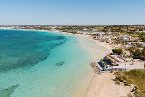 An aerial view of a beachside property with clear turquoise waters, a sandy beach, and a row of buildings with a row of palm trees in the background.