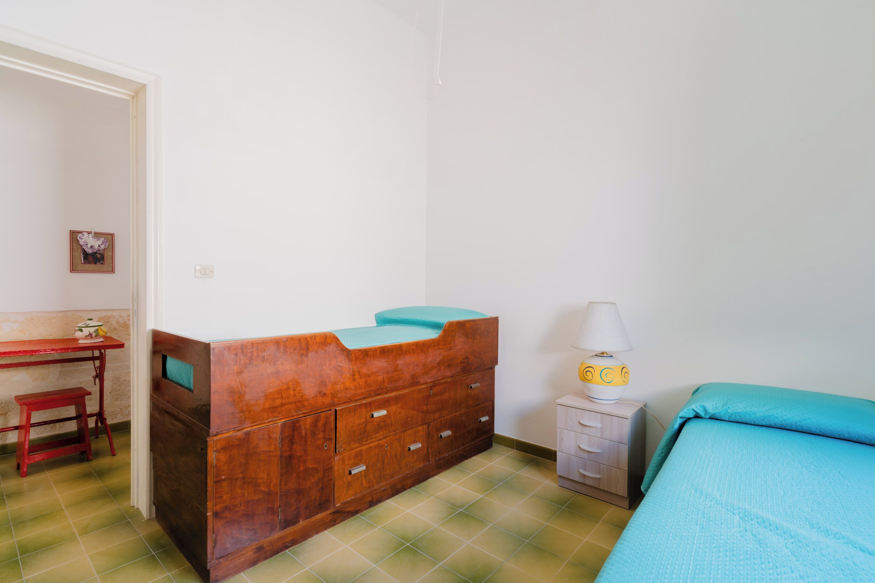 A small, simple room with a wooden dresser, a bed with a blue bedspread, a small table with a lamp, and a bench.
