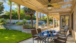 Eine überdachte Terrasse mit Esstisch, umgeben von Stühlen und Blick auf einen angelegten Garten mit Palmen.