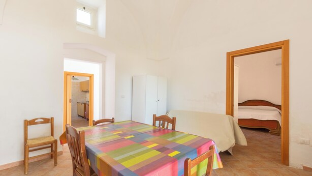 Ein Essbereich mit einem Tisch, der mit einer bunten Tischdecke bedeckt ist, Holzstühle und Blick auf ein Schlafzimmer mit Bett und Kleiderschrank.