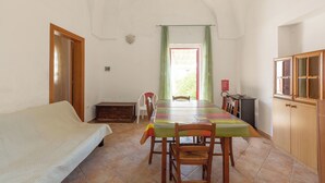 Ein Zimmer mit einem Bett, einem Esstisch mit Stühlen, einer kleinen Kommode und einem Fenster mit Vorhängen.