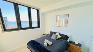 Une chambre avec une grande fenêtre, un lit recouvert d’une couvre-lit bleu foncé, deux tables de chevet et un tableau encadré accroché au mur.
