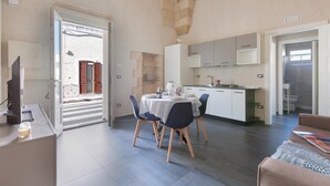 Een ruime woonkamer met een eettafel, stoelen en een bank. Er is een keuken met kastjes en een spoelbak. Een deur leidt naar een buitenruimte met uitzicht op een gebouw.