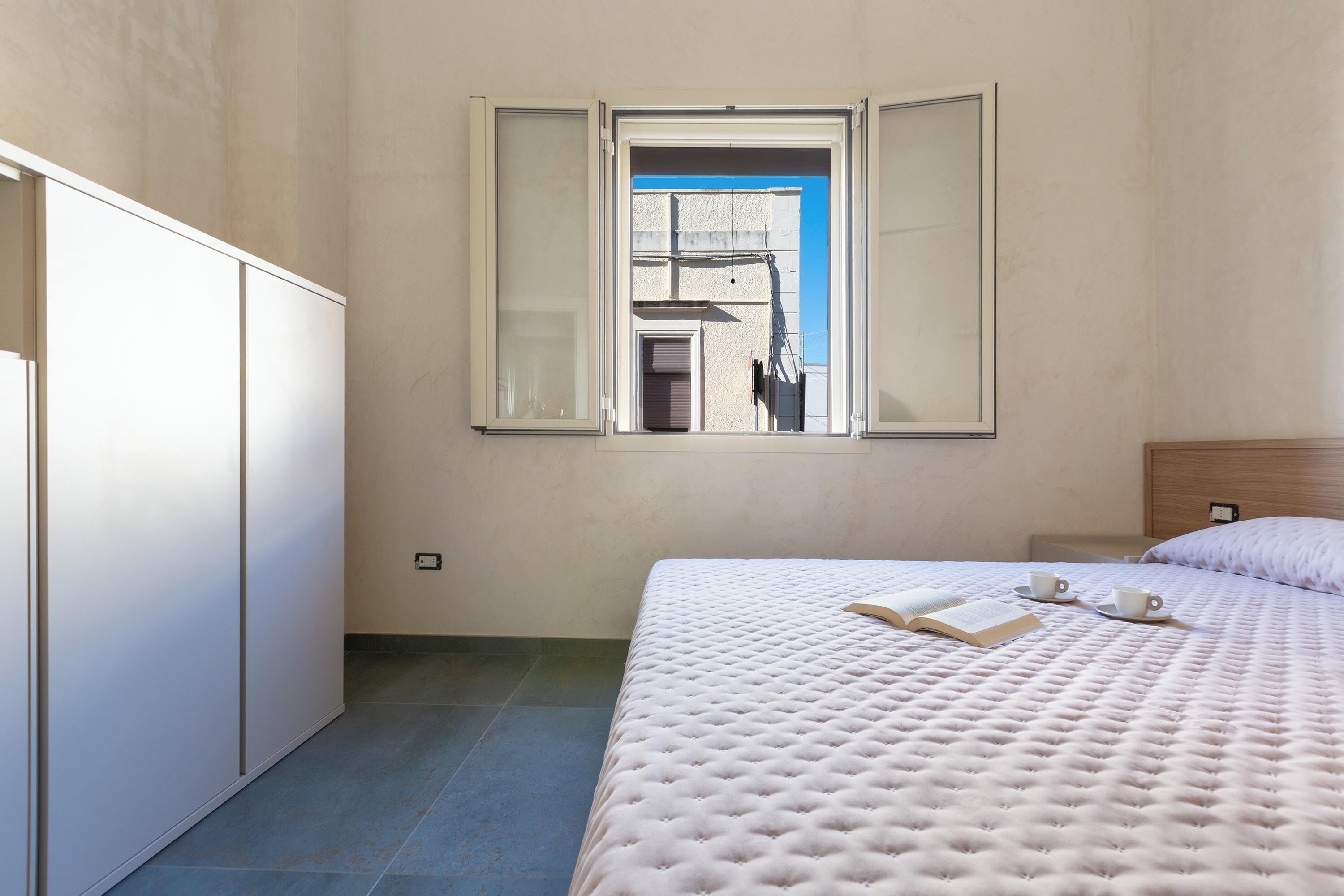 Une chambre avec un grand lit, une fenêtre dont le volet est ouvert, et une vue sur un bâtiment à l’extérieur.